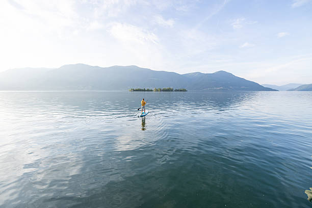 A man in a yellow hoodie paddleboarding on a calm lake. The peaceful water reflects surrounding mountains. Outdoor adventure and nature activity enthusiasts.