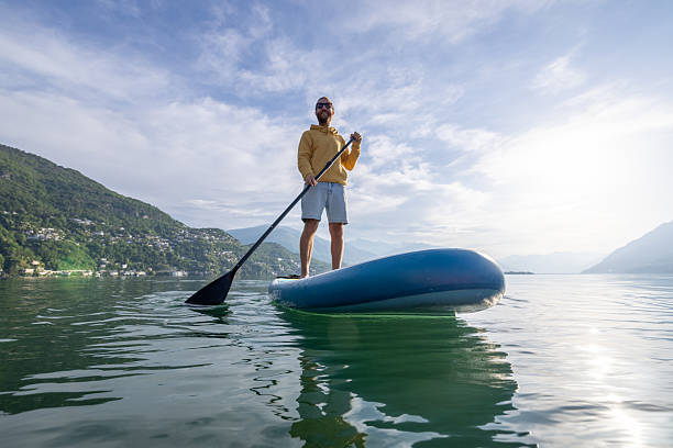Man in casual attire paddleboarding on calm lake, surrounded by mountains. Outdoor enthusiast enjoying peaceful nature. Active lifestyle and adventure on water.