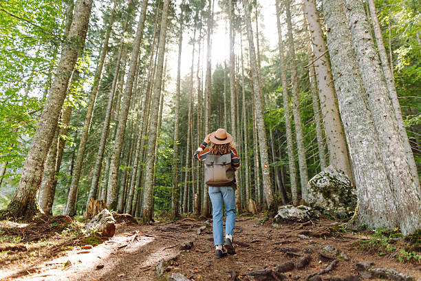 A female hiker with a backpack and wearing a hat walks along a lush forest path.