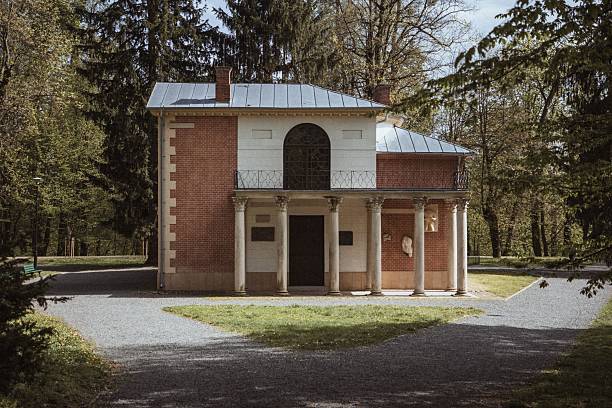 A historic building with columns set in a lush park under a partly cloudy sky, surrounded by tall trees, creating a serene atmosphere.
