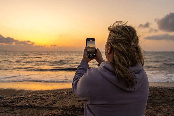 Middle-aged woman captures sunset on her smartphone while standing on beach. Back view. Soft golden light. Concept of love, serenity, emotional moments and meaningful solitude.