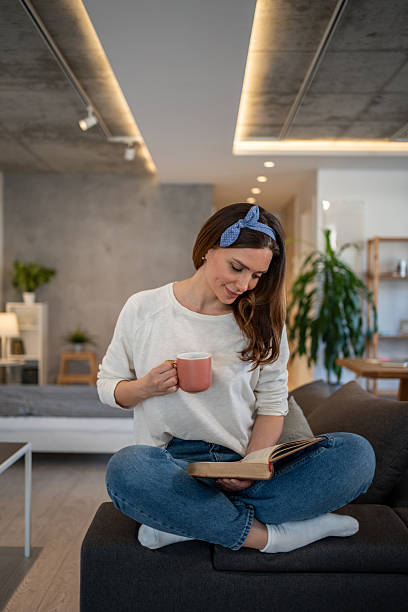 istock Young woman reading a book and drinking tea on sofa at home 2213721802