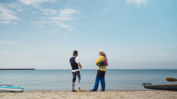 Two senior men with their sea sports equipment are chatting with each other by the sea at the beach. One man with an amputated leg has a stand-up paddle-board, and the other man has a kayak. They both enjoy sea sports.