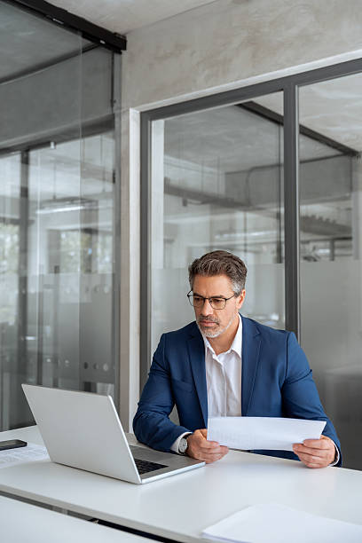 istock Entrepreneur manager businessman banking analyst doing paperwork analysing using pc. Busy latin mature business man working on laptop computer reading financial document report in office. Vertical 2213729817