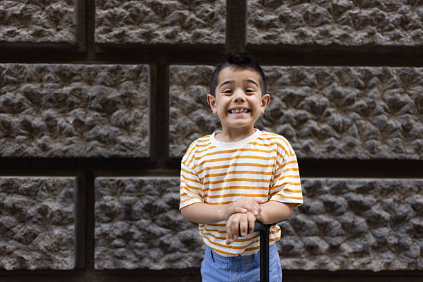 A cheerful young boy wearing a striped shirt and jeans stands joyfully against a brick wall, holding a handlebar, and smiling brightly at the camera.