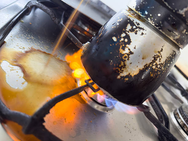 Dirty, burnt coffee pot is precariously perched on a gas stove, its spilled contents catching fire and creating a dangerous kitchen hazard