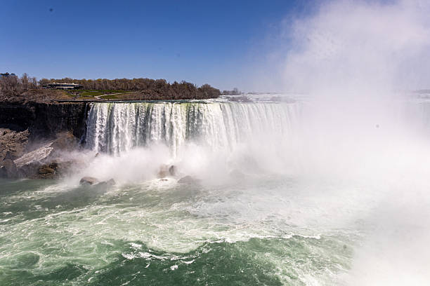 An awe-inspiring view of the iconic Horseshoe Falls, part of Niagara Falls, captured on a bright sunny day. Torrents of water plunge into the river below, creating a dramatic spray of mist that rises high into the air. The green-blue waters of the Niagara River add contrast to the power and beauty of this natural wonder. Ideal for themes such as travel, nature, landscapes, tourism, water power, and iconic destinations.