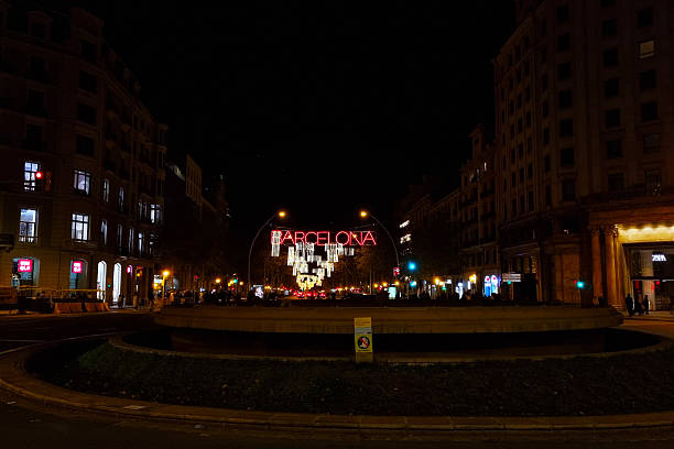 Barcelona, Spain, December 25, 2024: Nighttime view of a bustling barcelona intersection featuring a large illuminated barcelona sign. surrounded by historic buildings, the area is alive with vibrant lights and city energy, capturing the essence of barcelona s nightlife and cultural ambiance.