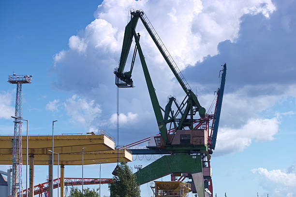 A large green crane is actively lifting a heavy structure at a busy construction site near the waterfront. The sunny weather features scattered clouds and clear visibility.