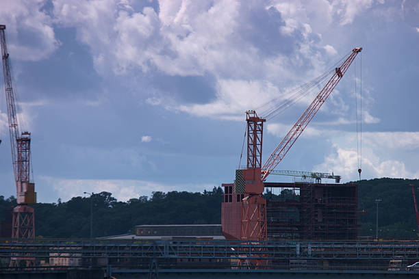 Cranes are actively lifting materials at a construction site located by the water. The skies are overcast, creating a dramatic backdrop. This scene captures industrial progress.
