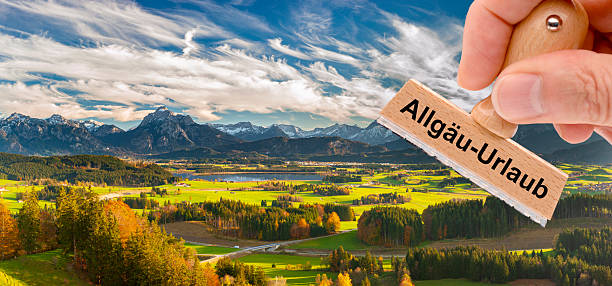 A wooden stamp in the hand is inscribed with the imprint Allgaeu holiday in front of the Alpine panorama in Bavaria - in German language Allgäu-Urlaub