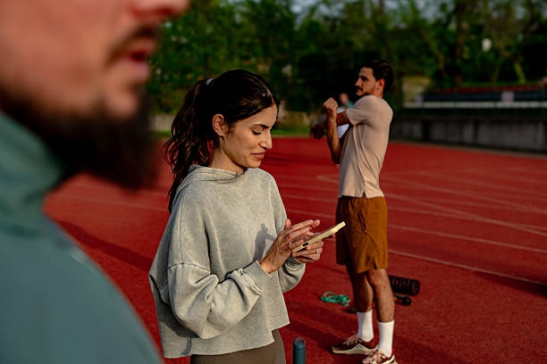 Young woman using phone on running track with athletes stretching