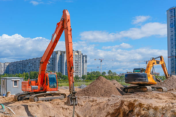Two large excavators is at work, digging into a mound of soil at a construction site. Tall residential buildings stand in the background under a clear sky.