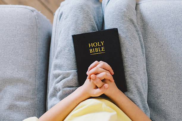 Close-up of a teenage girl reading a black book, with a rosary draped over her hand, suggesting a spiritual or religious context within a comfortable home setting.
