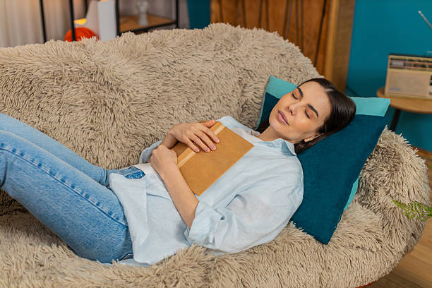 istock Young woman falling asleep lying on sofa peacefully holding open book tired after reading relaxing 2213772672