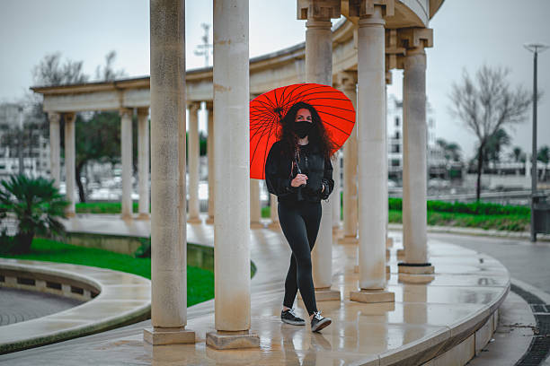 Young woman holding a bright red umbrella walks along a curved colonnade in rainy weather, contrasting her black outfit and face mask against classical architecture