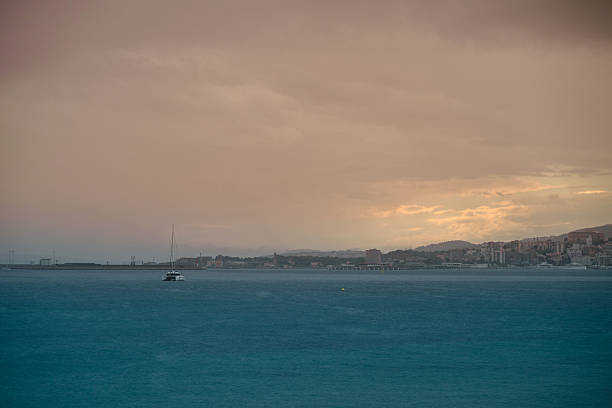 Serene view of a calm sea under a cloudy sunset sky, featuring a distant city skyline on the horizon and a solitary boat enhancing the tranquil atmosphere