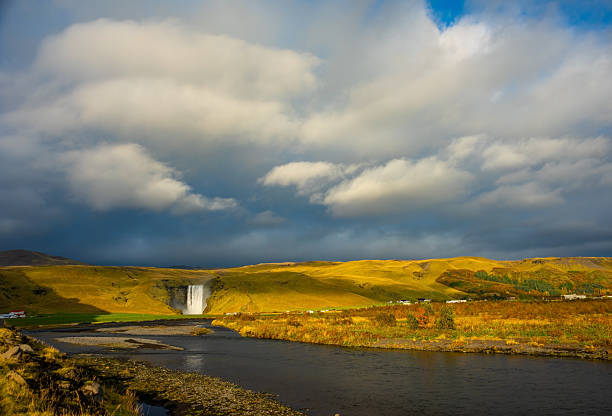 Skogafoss waterfall in Iceland cascades down a steep cliff, creating a powerful mist that forms a vibrant rainbow. The surrounding lush green landscape enhances its majestic beauty