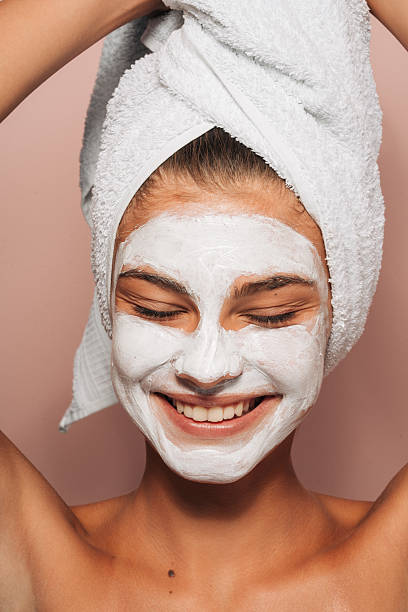 This is a close-up photo of a smiling woman having a clay mask treatment.She is smiling with towel on her head as she came out from the bathroom.  This is a studio shot