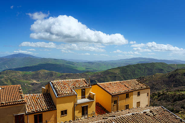 Charming Sicilian village houses with clay-tiled roofs overlooking lush green hills under a clear blue sky.