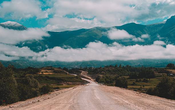 Scenic view of a winding dirt road leading through lush green fields towards majestic mountains under a partly cloudy sky.