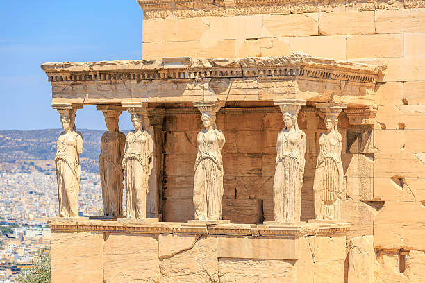 A group of statues are displayed on a building. The statues are of women and are arranged in a row. The building is located in a city and the statues are on a balcony, Acropolis in Athens