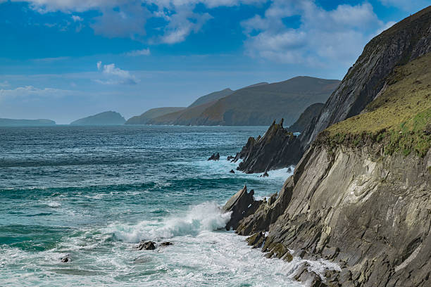 Waves crash against rugged cliffs under a bright blue sky, with distant islands visible across a vast expanse of ocean. The dramatic coastal landscape is enhanced by the contrasting colors of the deep green sea and the rocky terrain at Coumeenoole Beach, County Kerry, Ireland.
