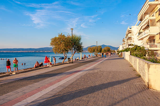 A beach scene with people walking along a sidewalk and a tree. Scene is relaxed and leisurely. August 8, 2024 Perea Greece