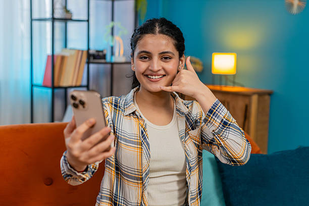 istock Indian woman sitting on sofa with smartphone making call me gesture smiling inviting communication 2213794299