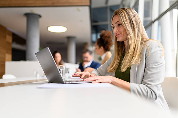 istock Focused blond businesswoman analyzing data over laptop while colleagues collaborating in the background in board room 2213799661