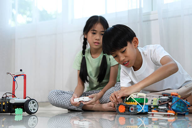 istock Asian children building and testing robotics on the floor at home. A boy works on assembling a robot car while a girl watches and controls another robot with a remote. STEM learning and hands-on education concept for kids. 2213800152