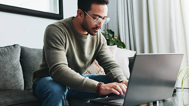 istock Typing, online and laptop with man on sofa for freelancer, networking and email response. Reading, communication and remote worker with person in living room of home for business proposal or research 2213829381