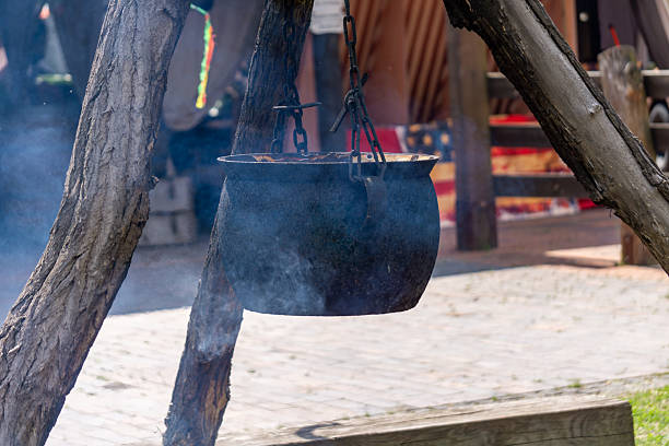 traditional cast iron cauldron hanging over an open fire, suspended by a rustic wooden tripod. Smoke rises from the pot, suggesting outdoor cooking in a historical or primitive setting. Perfect for themes related to history, survival, outdoor living, or traditional cuisine