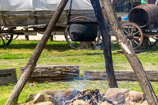 cast iron pot hanging over an open campfire in a rustic, Wild West-style encampment. In the background, a wooden barrel and a horse-drawn wagon add to the historical and outdoor atmosphere. Perfect image for illustrating themes of outdoor cooking