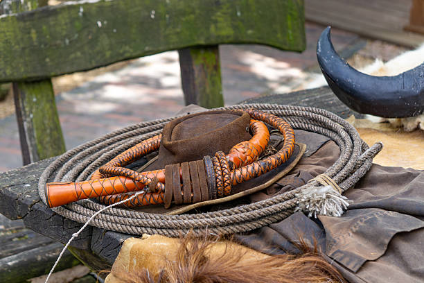 rustic composition featuring cowboy-themed accessories including a leather hat, braided whip, rope, and an animal horn, arranged on a weathered wooden bench. The scene evokes the atmosphere of the Wild West, ideal for illustrating themes related to western history, cowboy culture, or historical reenactments. Natural light and close-up detail enhance the authenticity.