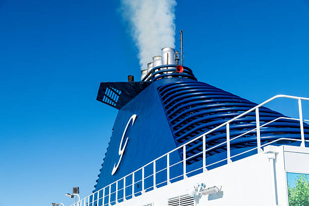 Barcelona, Catalonia - Spain - 01-10-2025: View from below of Grimaldi Cruise Roma ferry funnel with white smoke, metal railing, deck lights, and ventilation pipes against blue sky.