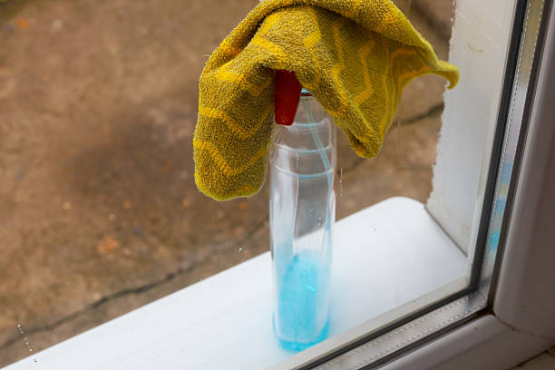 A plastic bottle with blue liquid and a yellow rag lie on a white windowsill by a window. Glass cleaning supplies, ready for use in the household.