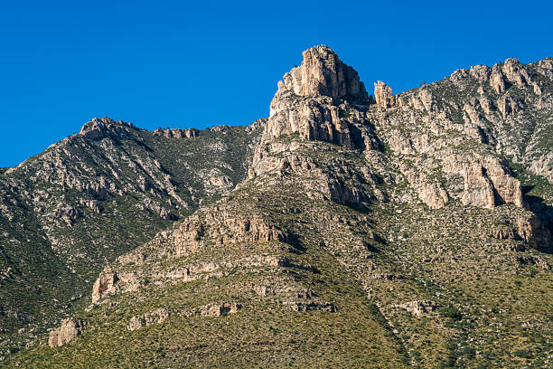 Rugged red sandstone cliffs rise steeply against a clear blue Nevada sky, their layered crimson and beige strata etched by eons of wind and erosion in the stark desert expanse.
