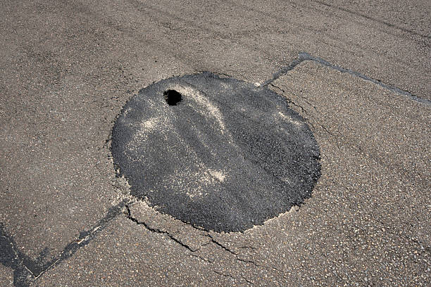 Potholes mar the road surface, collecting rainwater under an overcast sky in a city environment.