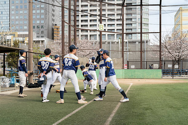 A local baseball team engages in teamwork and practice at an urban Tokyo field surrounded by buildings and cherry blossoms, emphasizing community spirit, friendship, and determination, set under Spring skies.