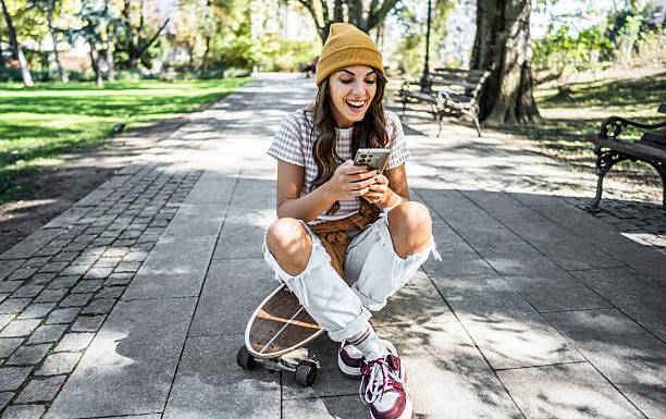 istock Young woman using mobile phone while sitting on skateboard in park 2213861152