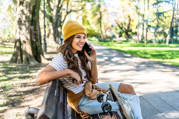 Smiling young woman talking on smartphone while sitting on bench in park with her skateboard next to her, enjoying sunny autumn day