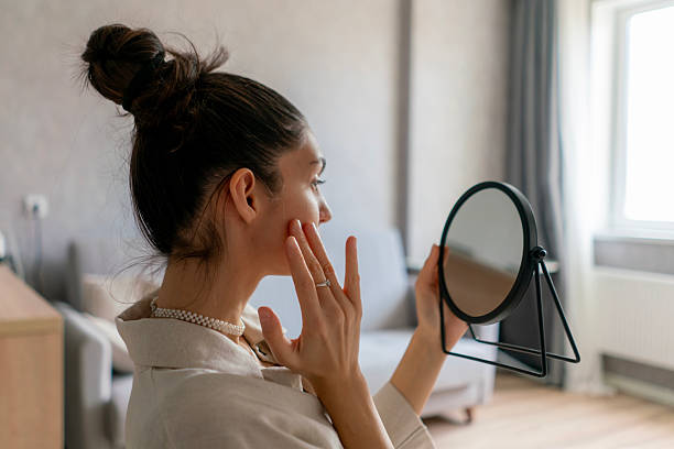istock A woman touches her cheek while looking into a small mirror, surrounded by a bright and inviting room with soft furnishings 2213869765