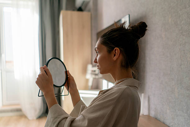 istock A woman touches her cheek while looking into a small mirror, surrounded by a bright and inviting room with soft furnishings 2213869782