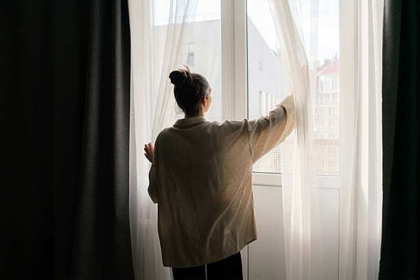 istock A woman stands by a window, pulling back curtains to let in natural light and brighten her living space 2213869915