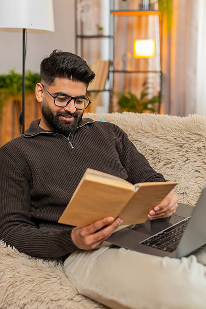 istock Indian man sitting on sofa with laptop and textbook, flipping pages while preparing for exam at home 2213877490