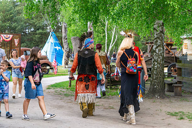 Józefów, Greater Poland Voivodeship, Poland - May 02, 2025 lively scene of park visitors and performers dressed in Native American-inspired regalia walking through a Western-style heritage village. Surrounded by rustic buildings, old vehicles, a tipi, and American flags, the image captures a vibrant, multicultural atmosphere blending education, entertainment, and family fun. Ideal for stock use in travel promotion, themed park events, or cultural experience advertising