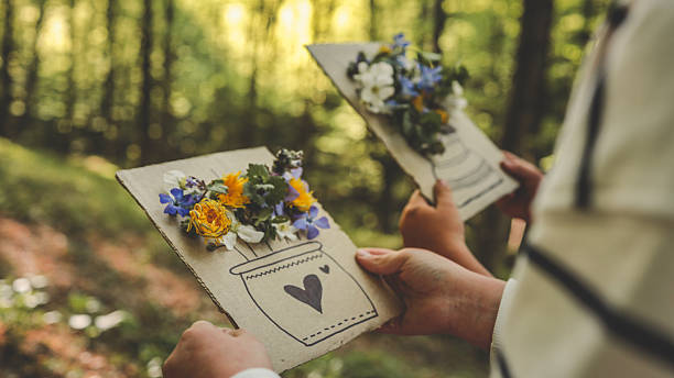 istock Two children holding handmade spring crafts created from real wildflowers on cardboard with vase drawings, standing in a sunlit forest during an outdoor creative activity 2213885487