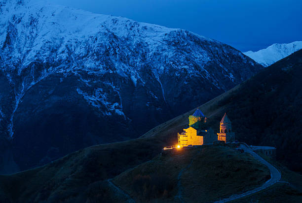 A captivating twilight scene captures the iconic Gergeti Trinity Church near Stepantsminda, Georgia, illuminated against the darkening sky and the imposing, snow-dusted Mount Kazbek in the background. The warm glow emanating from the church contrasts beautifully with the cool blues and whites of the surrounding mountains and the fading light, highlighting the spiritual and majestic presence of this landmark