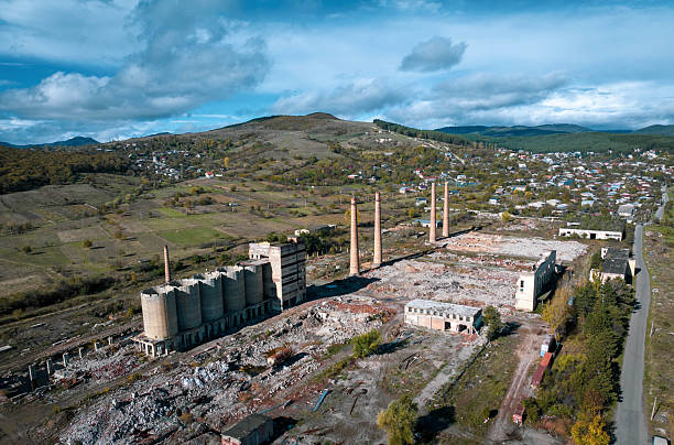 An aerial view captures the remnants of a large, seemingly abandoned industrial complex situated near Gori, Georgia. Concrete silos, dilapidated buildings, and towering smokestacks stand amidst a landscape showing signs of disuse and decay. The surrounding area includes sparse vegetation, a road, and a distant view of a town and rolling hills under a partly cloudy sky, hinting at a past industrial activity now overtaken by time.